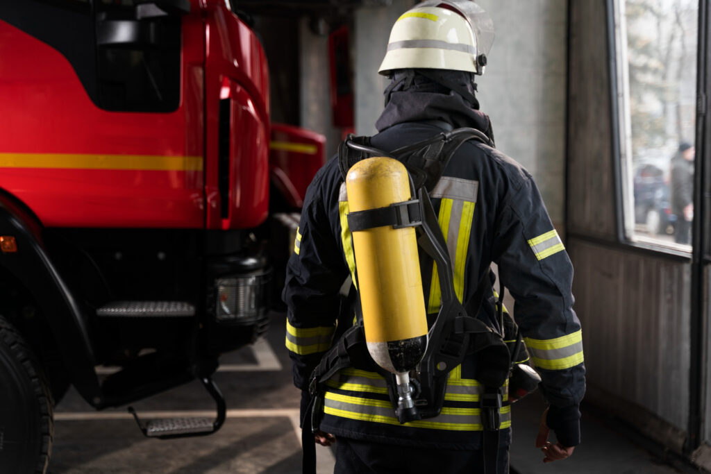Bombeiro com equipamento de proteção e cilindro de ar em ambiente de quartel.