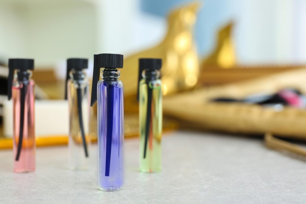 Quatro frascos pequenos de amostra de perfume, com liquidos nas cores rosa, transparente, azul e verde, alinhados sobre uma mesa clara